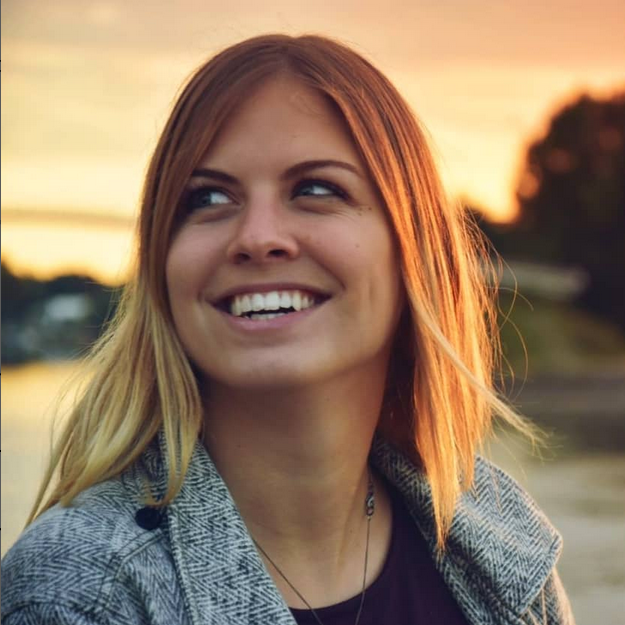 Das Bild zeigt eine junge Frau, die lächelnd in die Ferne schaut. Sie hat mittellanges, blondes Haar und trägt eine graue Jacke über einem dunklen Oberteil. Der Hintergrund zeigt eine unscharfe Landschaft mit Wasser und Bäumen, und der Himmel ist in warmen Orangetönen gefärbt, was auf einen Sonnenuntergang hinweist. Die Atmosphäre wirkt friedlich und harmonisch.